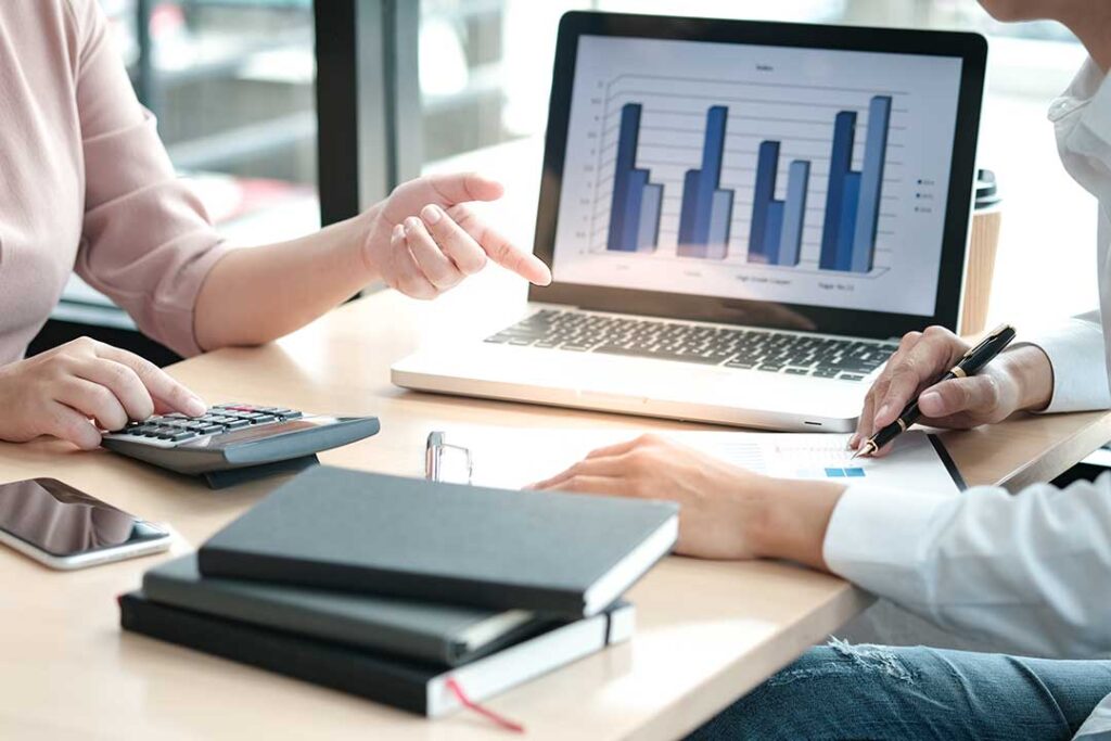 A person sits at a kitchen table reviewing documents with a laptop and calculator.